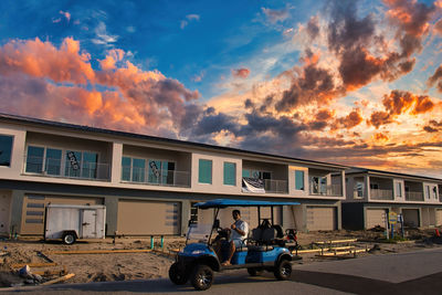 People on road by buildings against sky during sunset