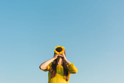 Low angle view of woman covering face with sunflower against clear blue sky