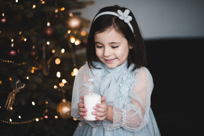 Cute girl holding candle sitting on sofa by illuminated christmas tree