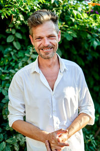 Happy middle aged businessman in white shirt clasping hands and looking at camera while standing near lush bushes on summer day in garden