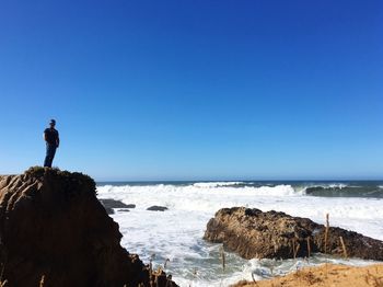 Man standing on rock by sea against clear blue sky