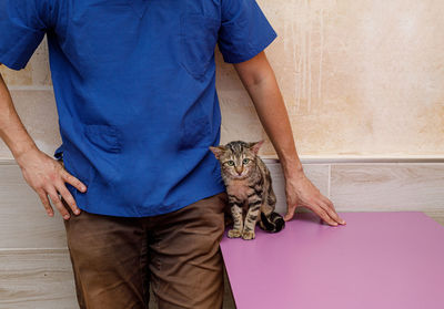 Crop male vet doctor in uniform standing against wall near table with curious scared cat and looking at each other in clinic