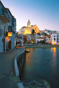 Illuminated street by buildings against sky at dusk