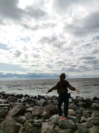 Full length rear view of woman standing on rock at beach against sky