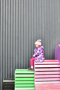 Side view of girl in warm clothing looking away while sitting on bench against wall