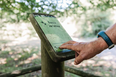 Cropped anonymous blind elderly traveler touching and reading braille on signboard while visiting park on summer weekend day