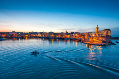 Illuminated buildings by sea against sky at sunset