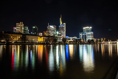 Illuminated buildings by river against sky at night