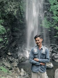 Portrait of young man standing against waterfall in forest