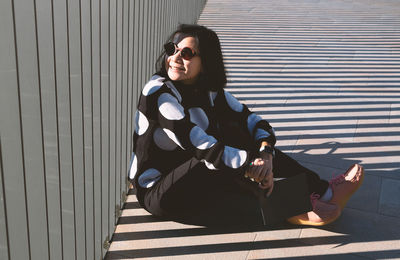 Side view of woman wearing sunglasses sitting on footpath