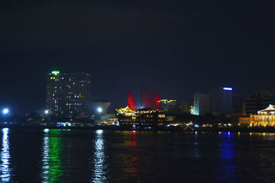 Illuminated buildings by river against sky at night