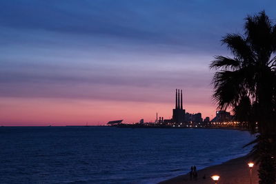 Silhouette buildings by sea against sky at sunset