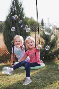 Portrait of smiling siblings gesturing towards bubbles while sitting on swing