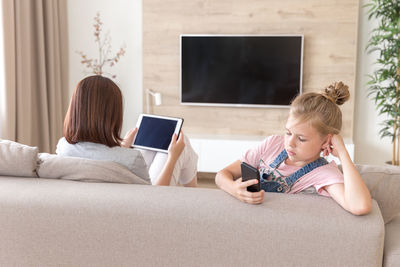 Woman holding digital tablet while sitting by daughter on sofa at home