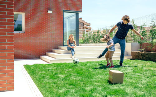 Mother sitting while father and son playing soccer in lawn