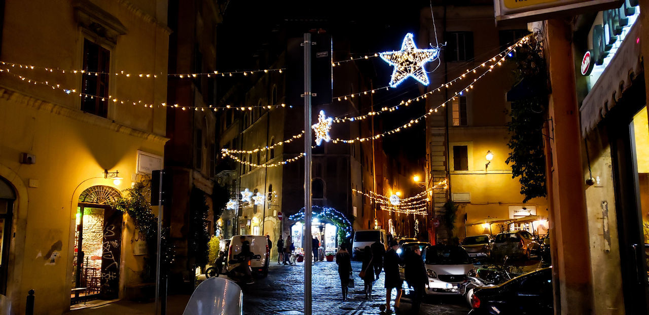 PEOPLE WALKING ON ILLUMINATED STREET AMIDST BUILDINGS AT NIGHT