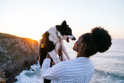 Side view of african american female owner holding cute happy border collie dog while enjoying time together near waving sea at sunset