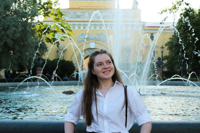 Smiling young woman looking away while standing against fountain and building