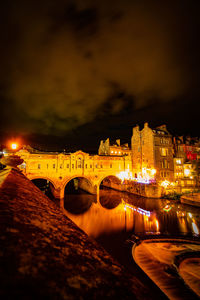 Illuminated bridge over river by buildings against sky at night