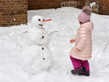 Little girl dressed in pink winter attire is putting the finishing touches on a snowman