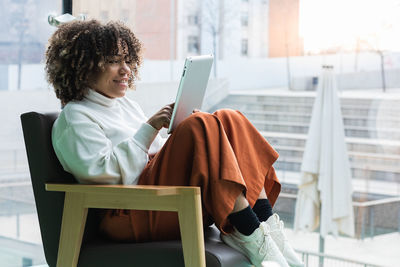 Side view of content african american female sitting in comfortable armchair and browsing tablet while entertaining during weekend at home