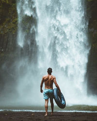 Rear view of shirtless man standing against waterfall