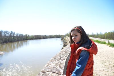 Side view of girl in warm clothing standing on footpath by river against clear sky during sunny day