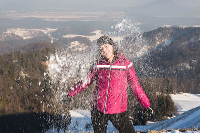 Portrait of smiling woman throwing snow standing against mountain range