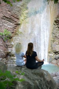 Rear view of woman sitting on rock against waterfall