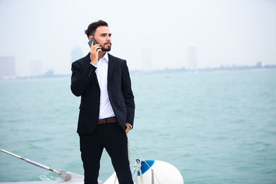 Businessman talking on phone while standing on boat in sea against clear sky