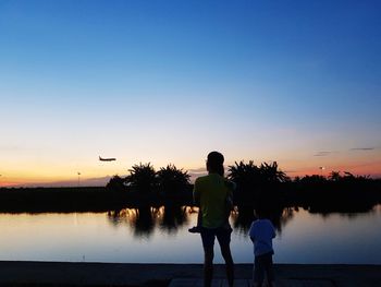 Rear view of family standing by lake against sky during sunset