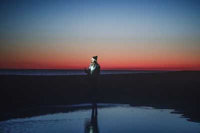 Woman using mobile phone while standing on beach against sky during sunset