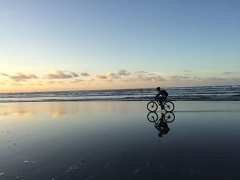 Side view of man riding cycling on sea shore against sky during sunset