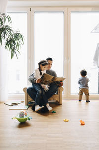 Full length of father reading book to daughter while son playing by window at home