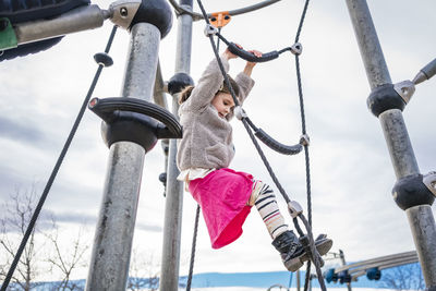 Low angle view of girl climbing on rope against cloudy sky at playground