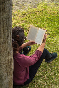 High angle view of woman reading book by tree trunk