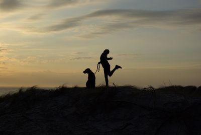 Silhouette woman with dog standing on land against sky during sunset