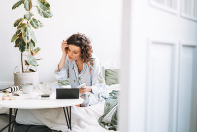 Young brunette woman in blue pajamas applies makeup with mobile phone sitting on bed at home