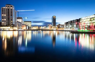 Illuminated buildings by river against blue sky at night