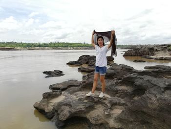Full length portrait of woman standing on rock at river against sky