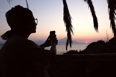 Silhouette man photographing by sea against sky during sunset