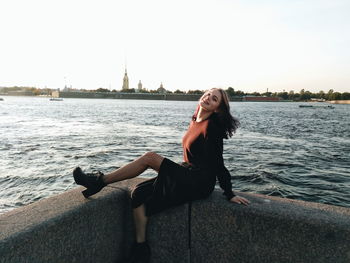 Full length of young woman sitting by sea against sky