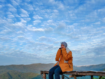 Young woman in warm clothing sitting on observation point against cloudy sky