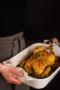 Anonymous cook in apron standing with baked chicken with rosemary on black background in studio
