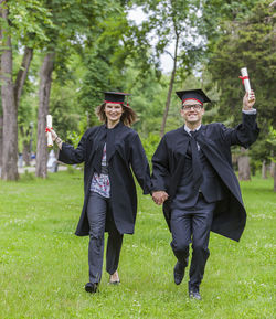 Full length portrait of couple wearing graduation gowns holding hands on field