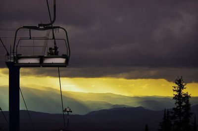 Ski lift by mountains against cloudy sky during sunset