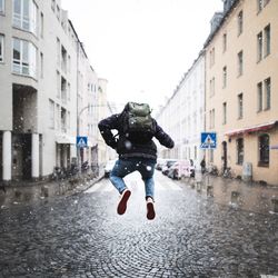 Full length rear view of man jumping on street in city against sky