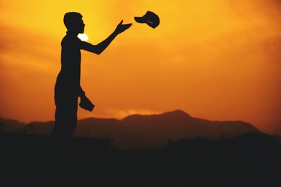 Silhouette man throwing cap against sky at sunset