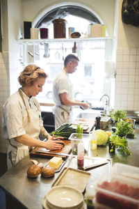 Female chef is cutting vegetables on board by colleague working at counter in restaurant kitchen