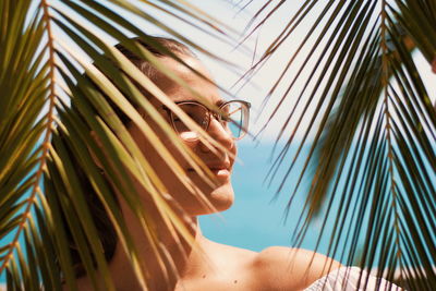Close-up of woman wearing eyeglasses seen through palm leaves at beach